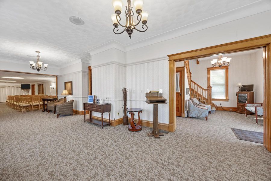 Spacious carpeted living room with chandeliers, white walls, and a doorway to a bedroom.