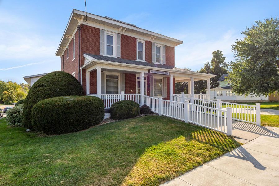Two-story red brick house with white trim, porch, and white fence on a sunny lawn