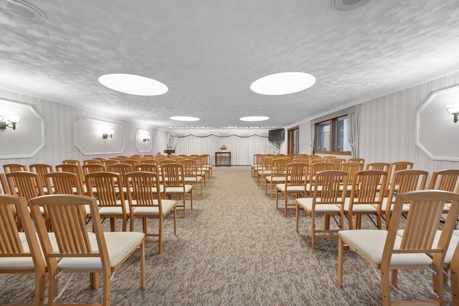 Empty conference room with rows of wooden chairs facing a front podium, carpeted floor, and bright ceiling lights