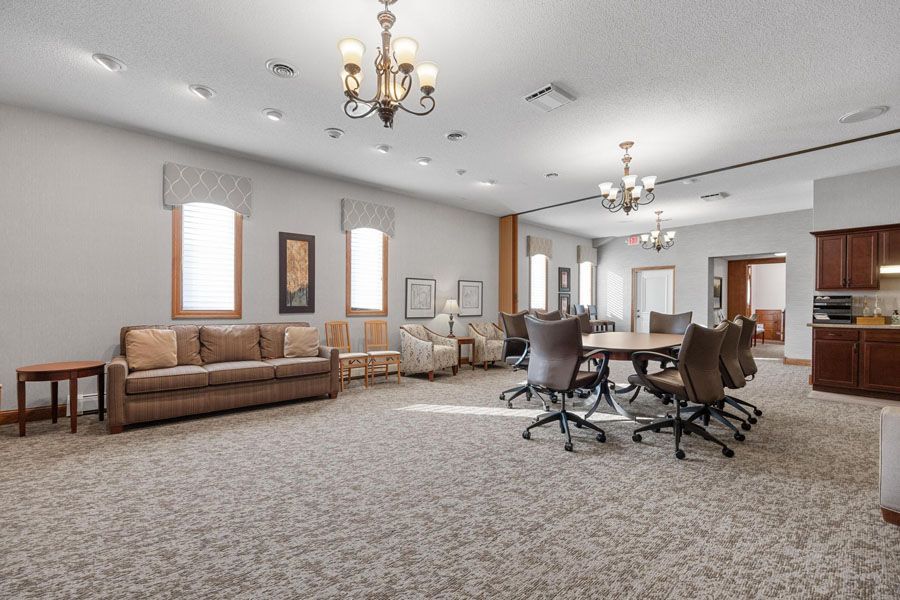 Spacious carpeted living room with sofas, chairs, table, chandeliers, and kitchen area in the background