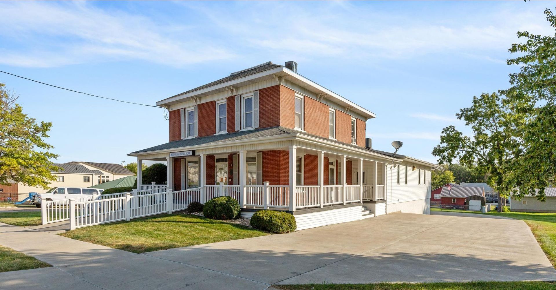Two-story red brick house with white trim, wraparound porch, and fenced yard on a sunny day