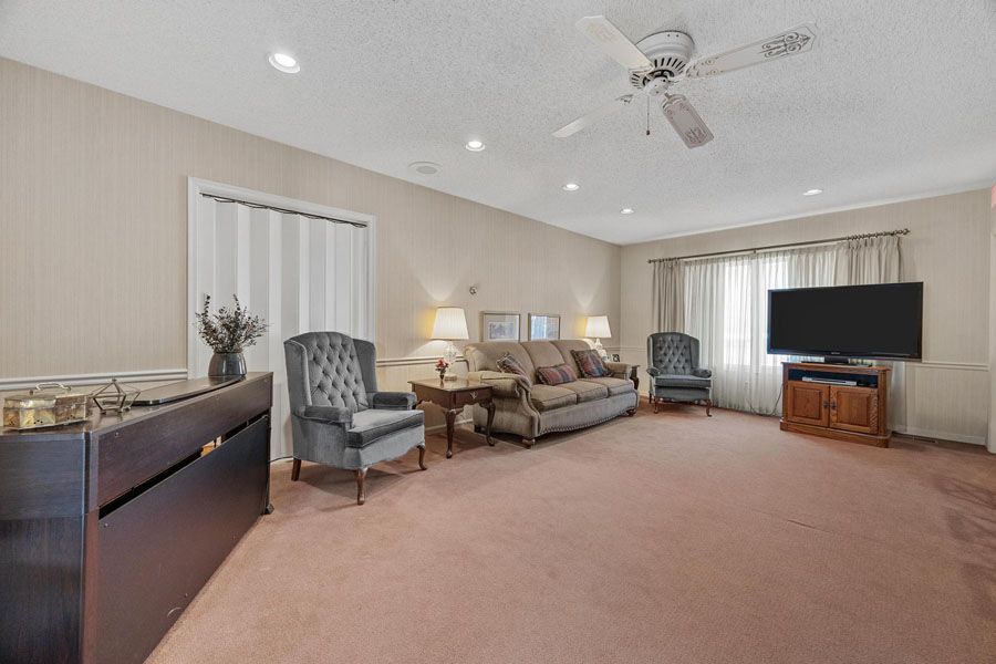 Spacious beige living room with sofa, armchairs, ceiling fan, and TV on a wooden stand.
