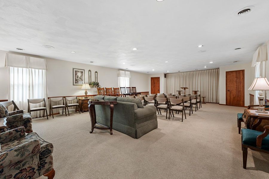 Spacious lounge with rows of chairs, a central couch, beige carpet, and large curtained windows