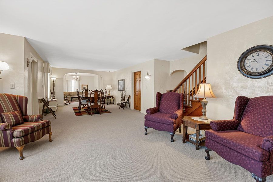 Spacious beige living room with purple chairs, dining table, staircase, and wall clock