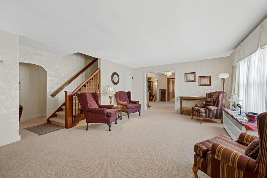 Bright living room with beige carpet, armchairs, staircase, and large windows with sheer curtains.