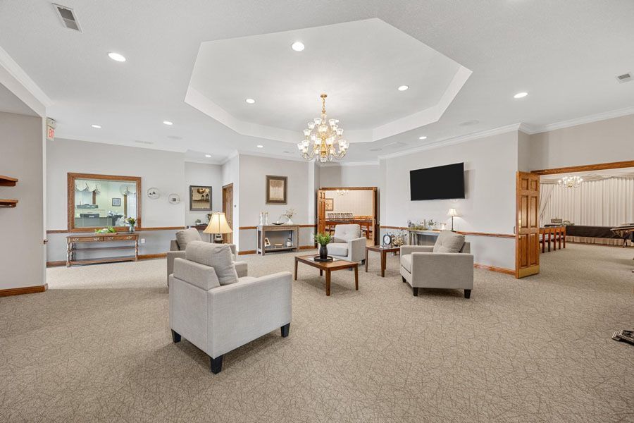 Spacious carpeted lobby with gray chairs, chandelier, wall-mounted TV, and wooden reception area