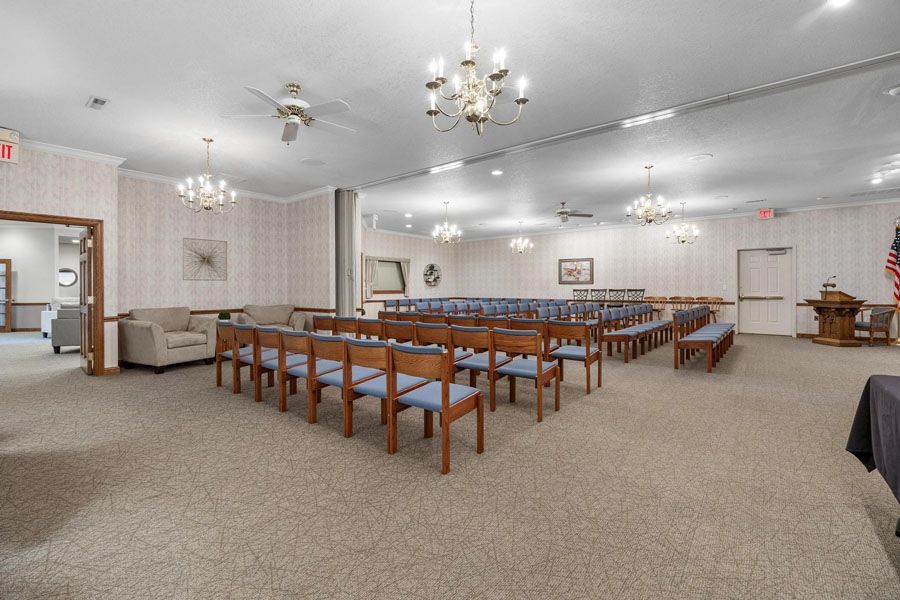Large hall with rows of wooden benches, chandeliers, and gray carpet in a bright, empty room