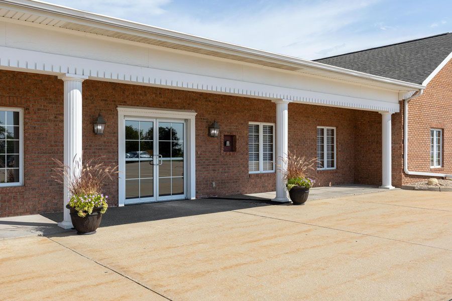 Brick building entrance with glass double doors, white columns, and potted plants on a concrete driveway.