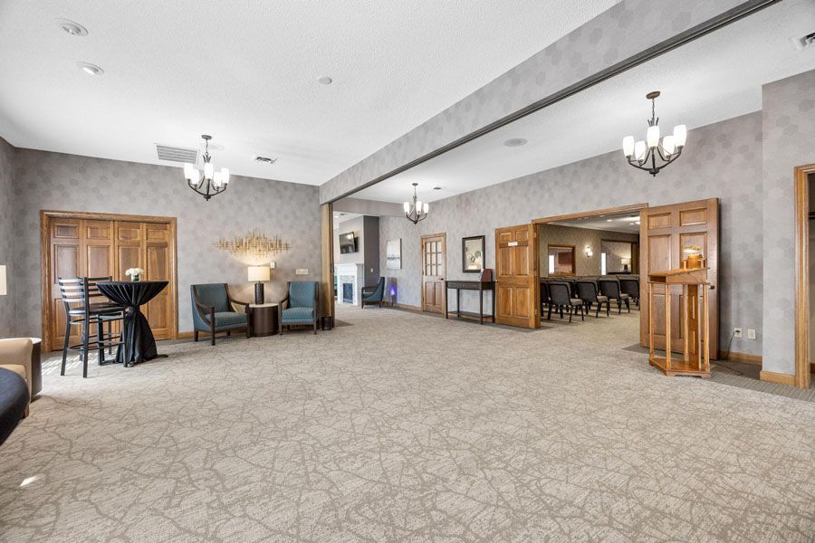Spacious beige banquet hall with carpet, chandeliers, wooden doors, and a bar area in back