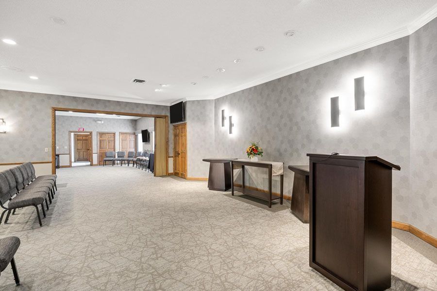 Wide, empty event hall with gray walls, carpeted floor, podiums, wall lights, and chairs along the side.