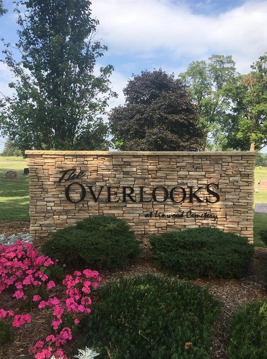 Stone entrance sign reading “The Overlooks” amid shrubs, pink flowers, and trees under a blue sky.