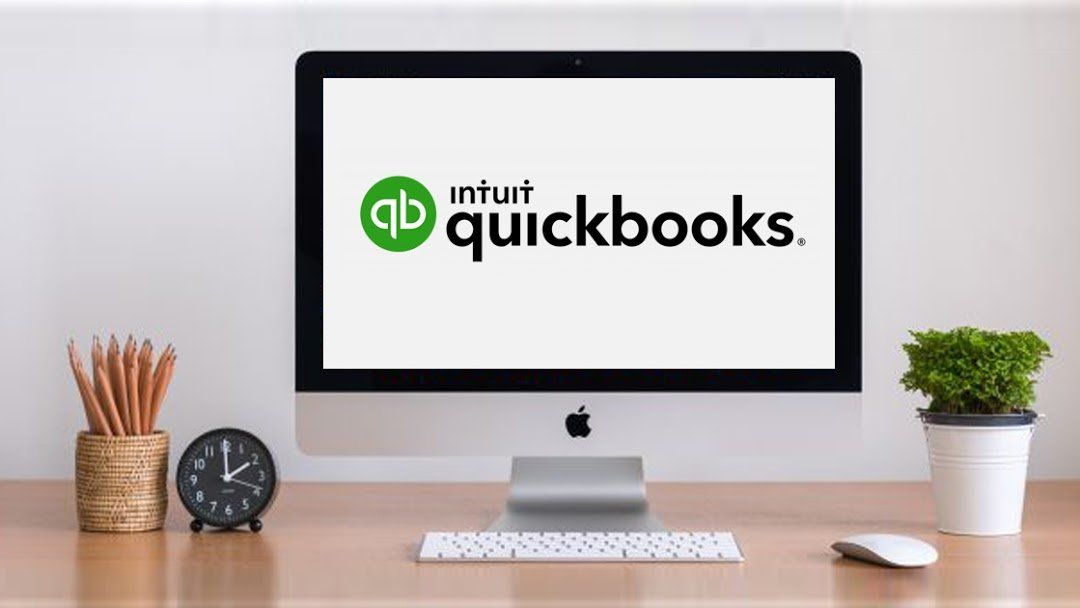 Desktop computer displaying the Intuit QuickBooks logo. Desk includes a pencil holder, clock, and small plant.