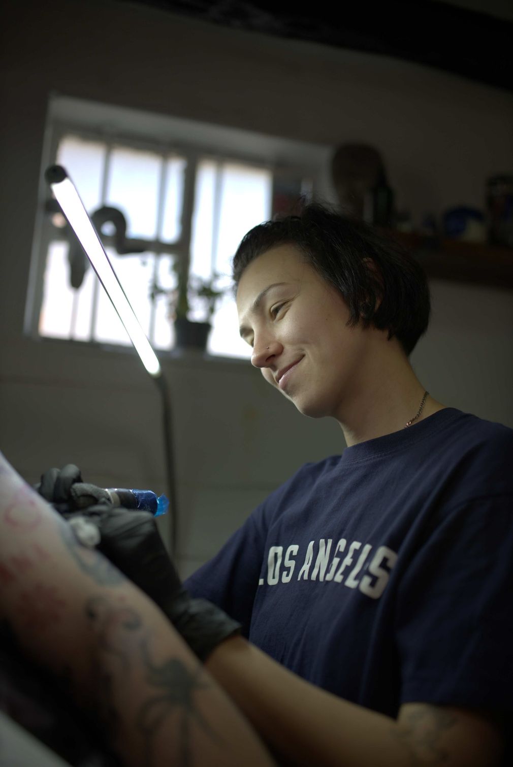 Tattoo artist with neck and arm tattoos sitting in a studio with supplies and artwork in the background.