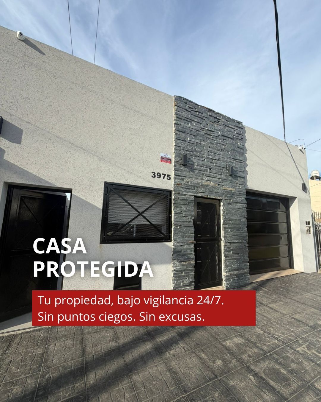 Casa con fachada blanca y muro de piedra; letrero rojo “CASA PROTEGIDA” sobre entrada y portón.