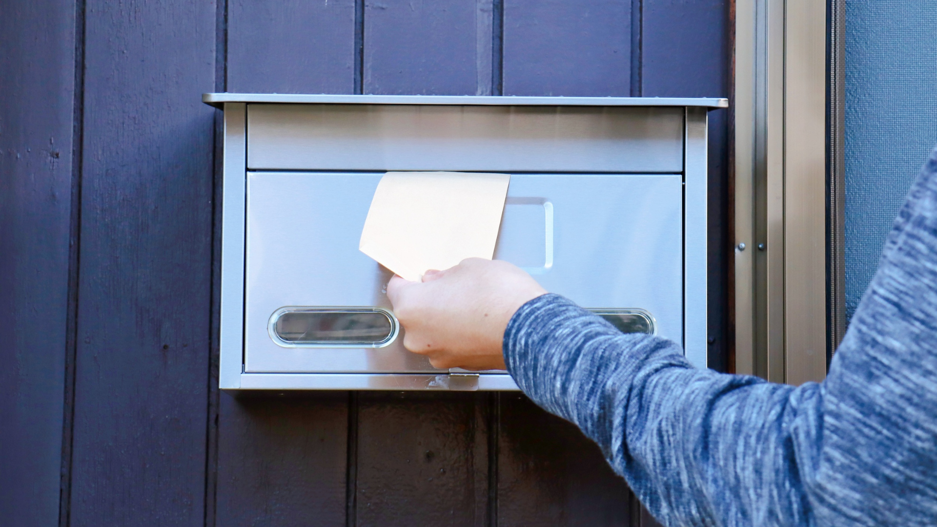 Person's hand placing a piece of paper into a silver mailbox mounted on a dark blue wooden door.