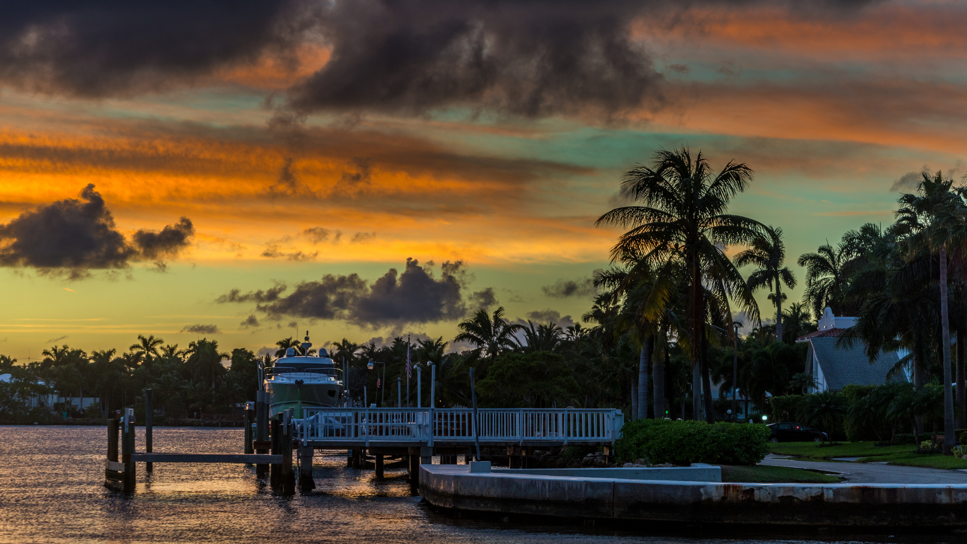 Sunset over water with dock, boat, palm trees, and vibrant orange and dark cloud sky.