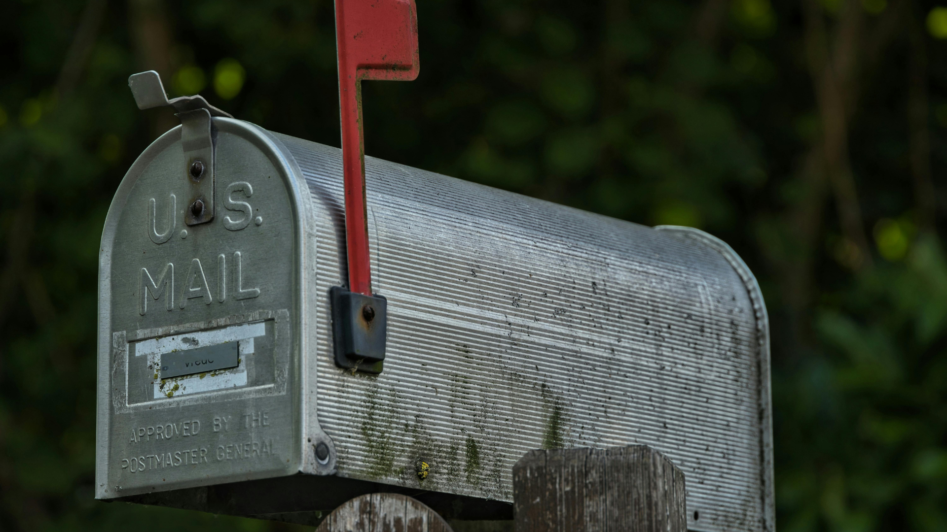 Silver U.S. Mail mailbox with red flag up, mounted on a wooden post, blurred green background.