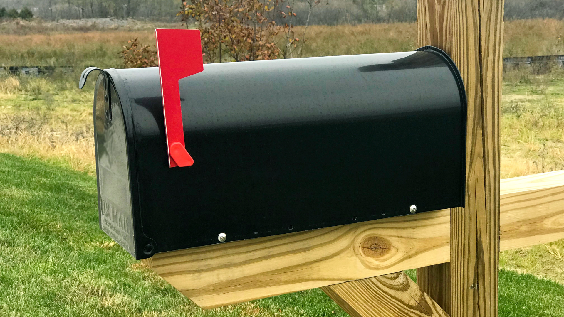 Black mailbox with red flag on wooden post, set in a grassy field.