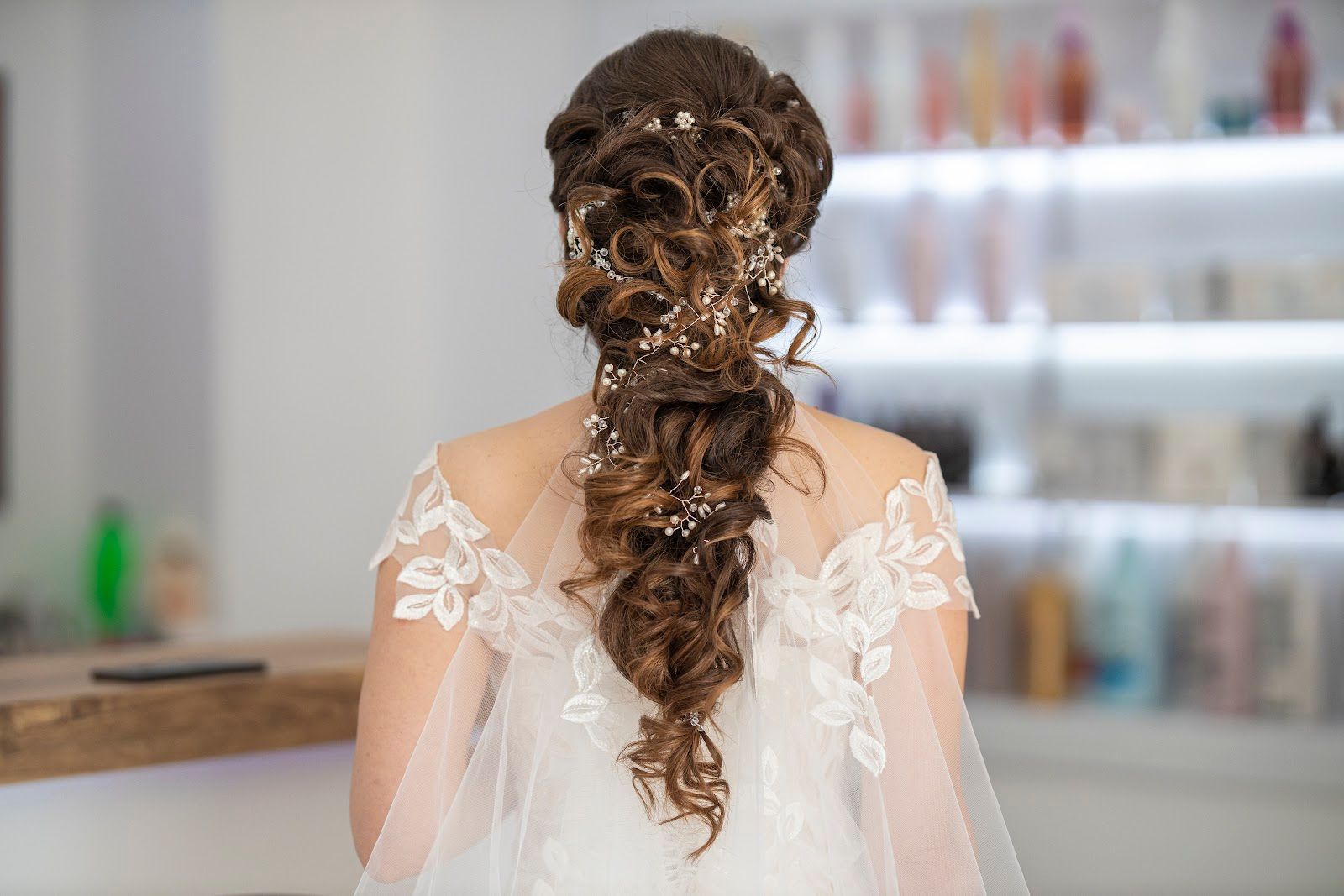 Woman with braided updo in a white lace dress, seen from behind in a salon.