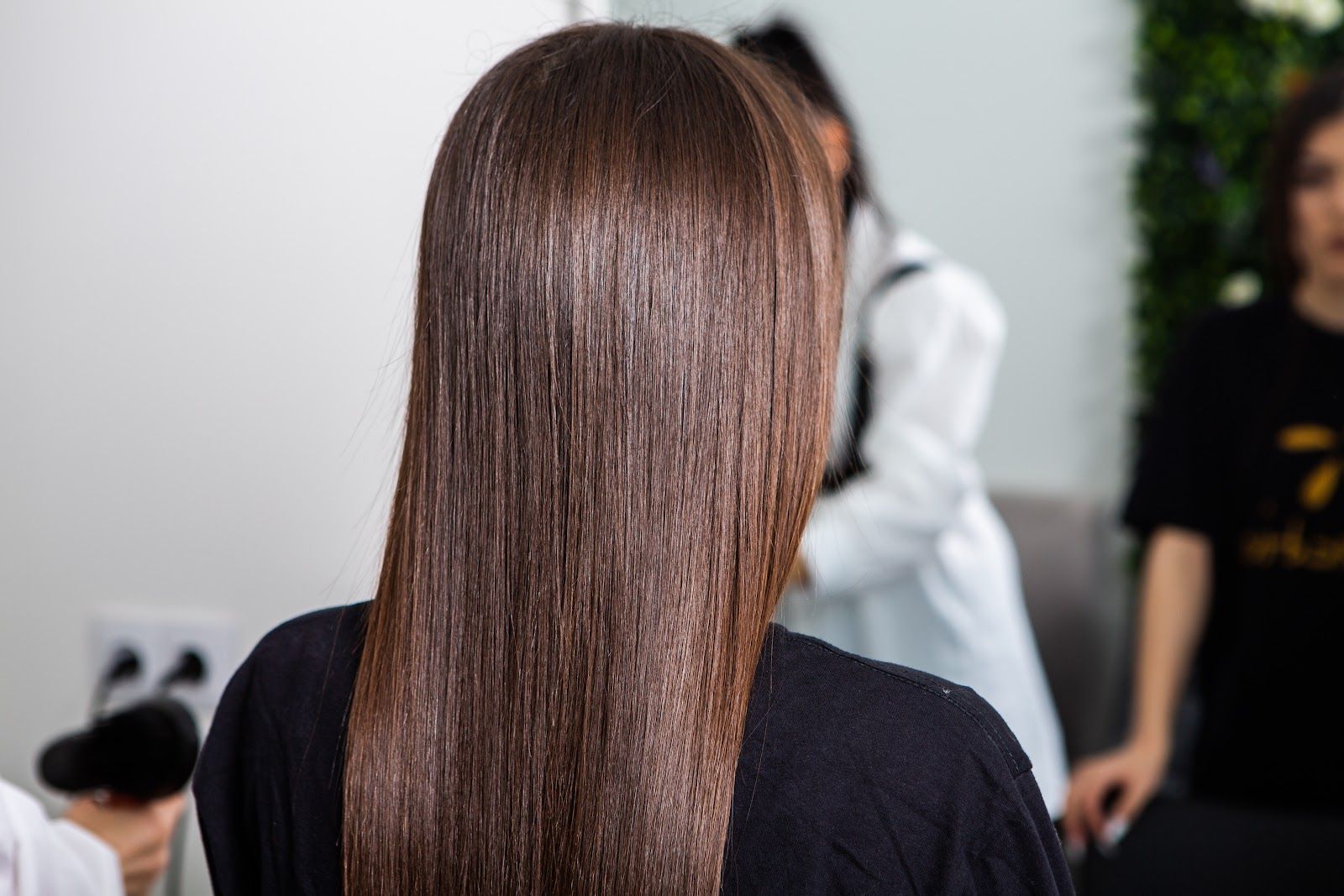 Woman with long brown hair standing in a salon, viewed from behind, with people blurred in the background