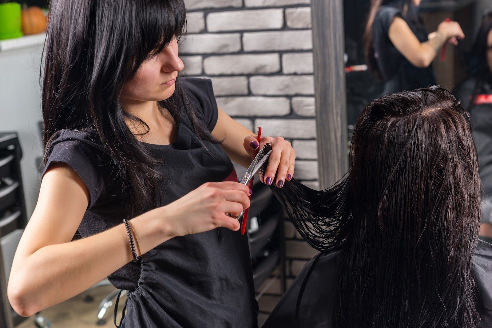 Stylist cutting a client’s long dark hair in a salon, with hair clip and brick wall background