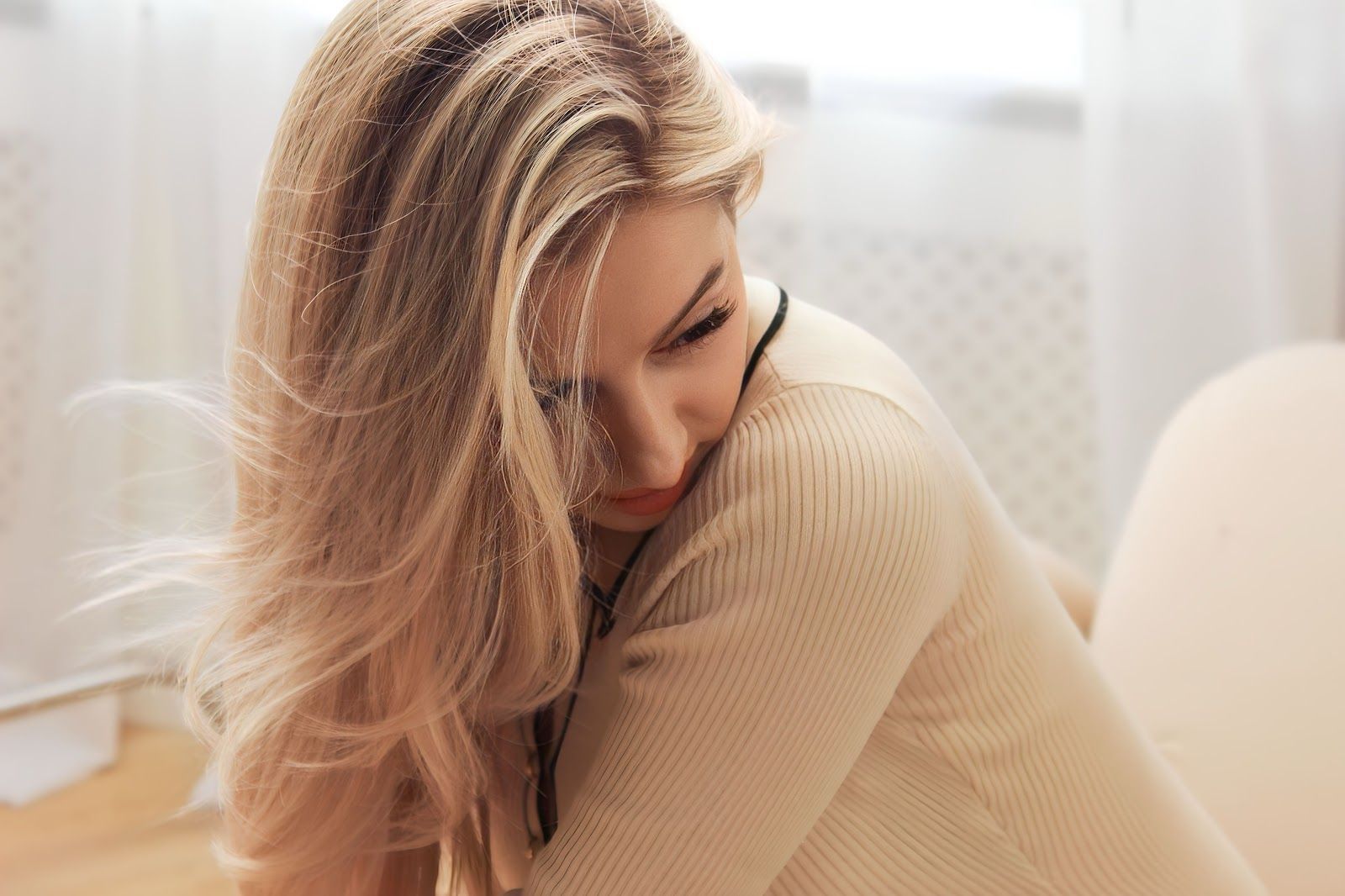 Woman with long blonde hair sitting indoors in a beige sweater, looking down thoughtfully