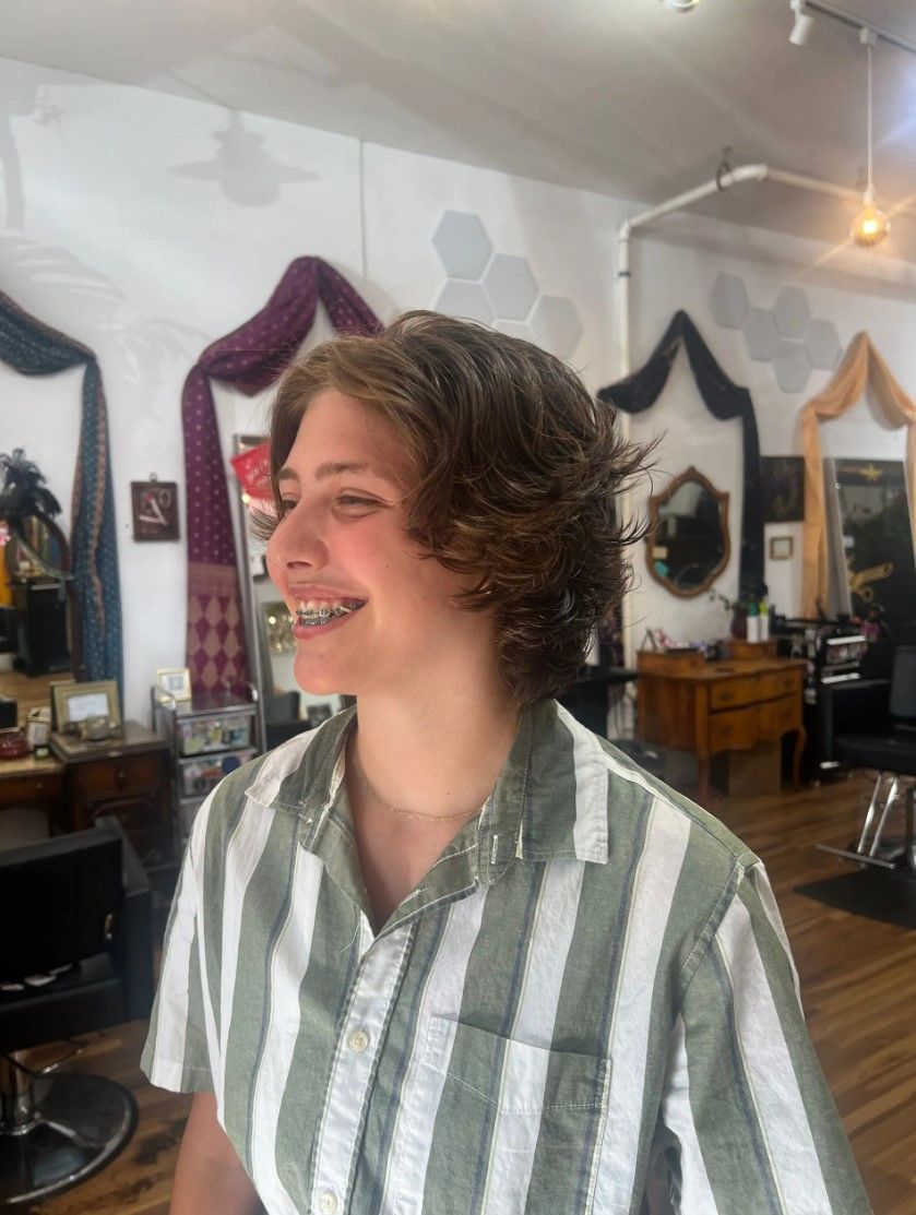 Person smiling in striped shirt at a salon, dark hair, braces. Background has decor and mirror.