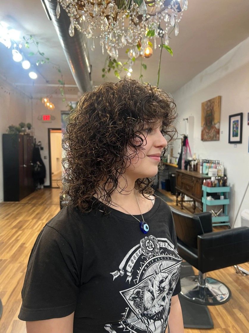 Woman with curly brown hair, wearing black shirt, stands in a salon.