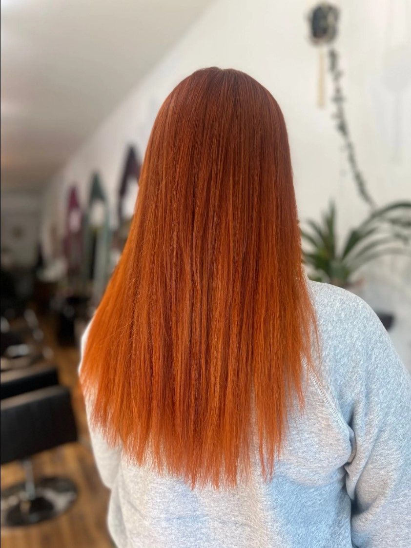 Woman with long, straight, vibrant orange hair; viewed from the back. Inside a salon.