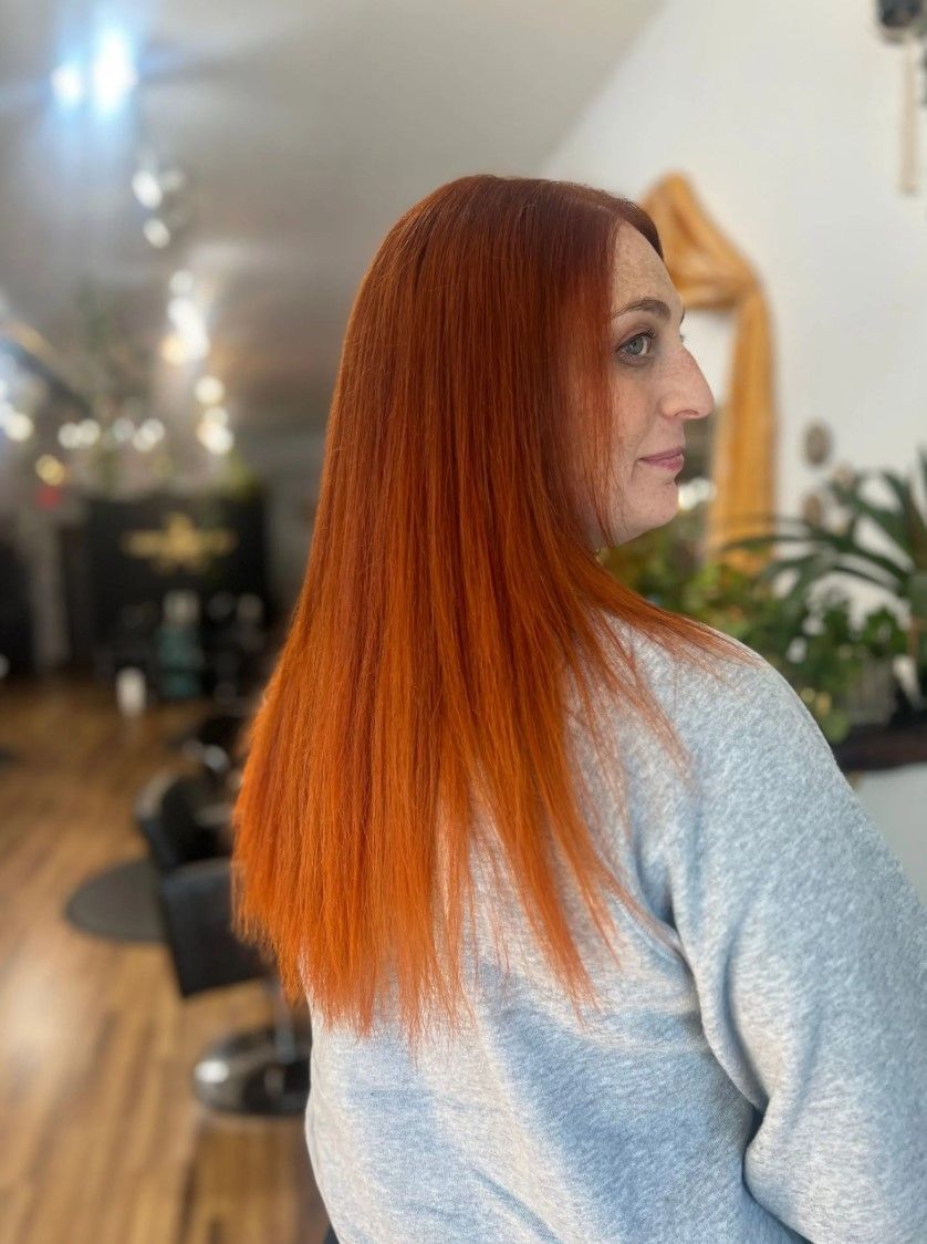 Woman with straight, vibrant red hair, in a salon, wearing a gray sweater.