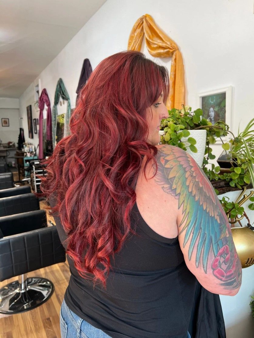 Woman with long, wavy red hair and colorful wing tattoo, standing in a salon.
