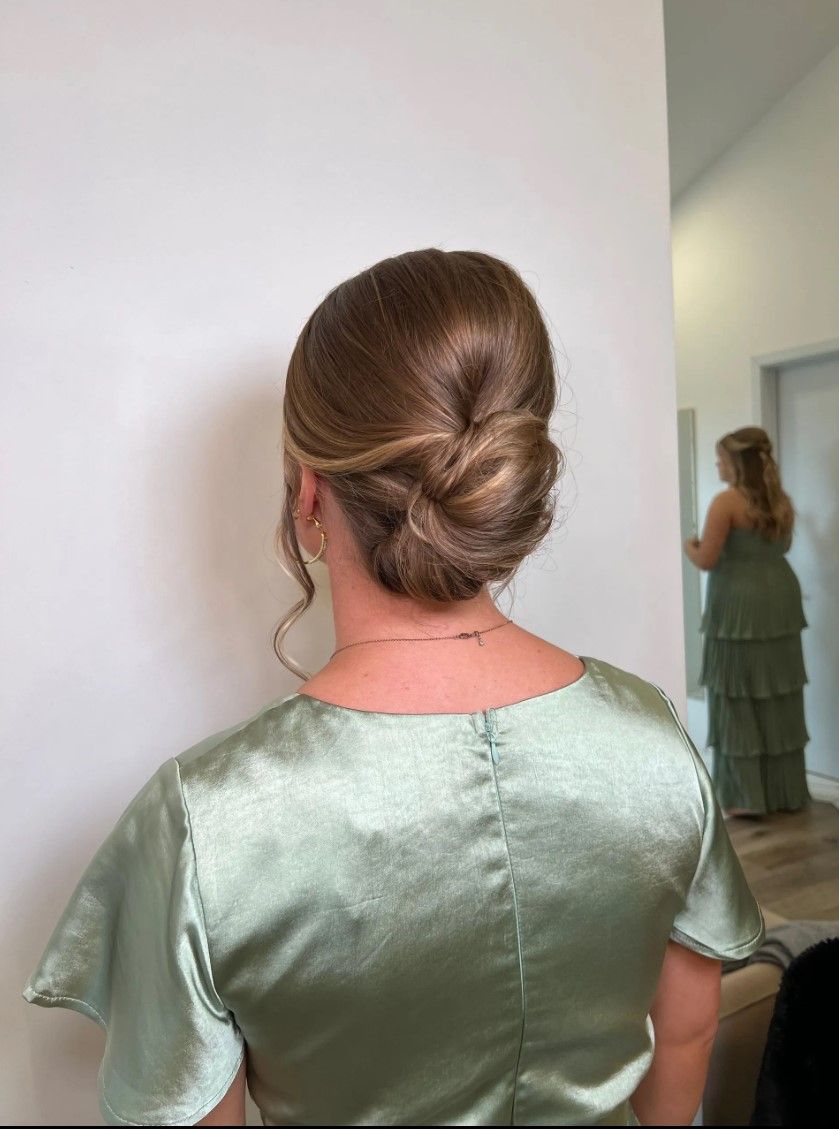 Woman with a light brown updo hairstyle, wearing a sage green satin dress, standing in front of a mirror.