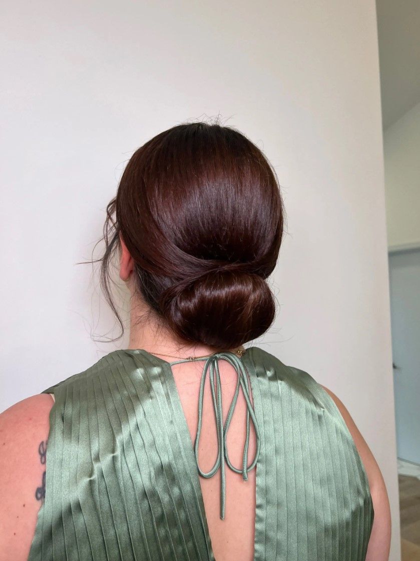 Woman with dark brown hair in a low bun, wearing a green satin top, against a white wall.