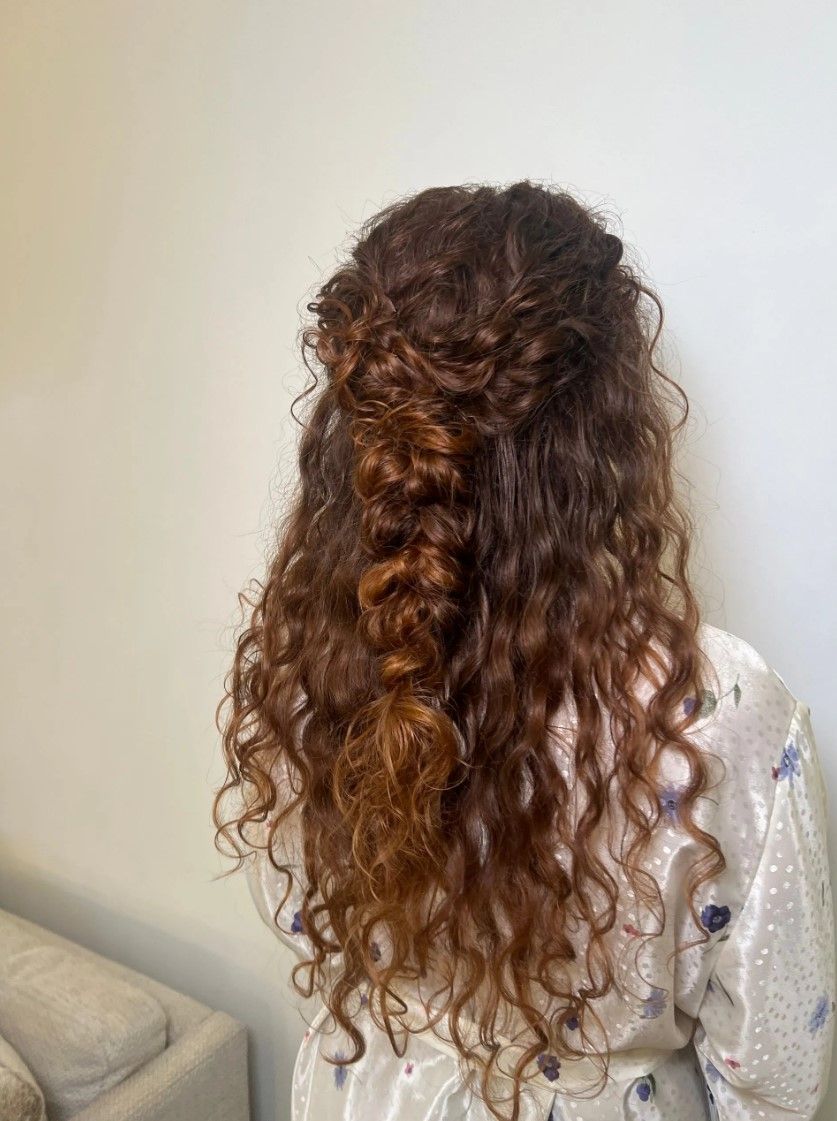 Woman with curly, reddish-brown hair styled in a half-up braided hairstyle, against a light-colored wall.