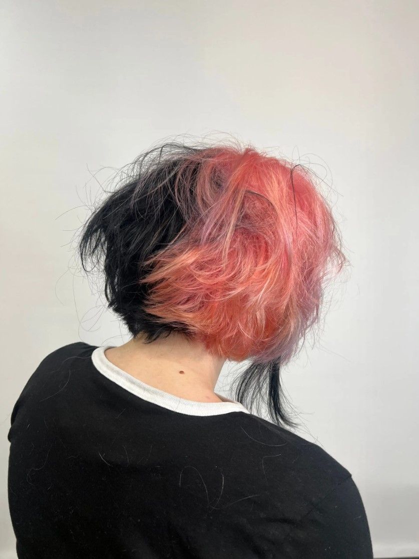 Person with short, two-toned hair: black on the left, pink and red on the right; wearing a black shirt.