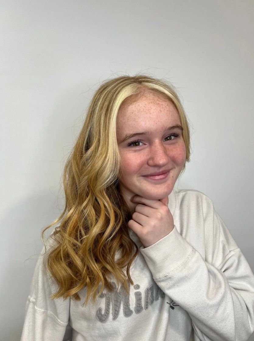 Blonde-haired person smiles, hand to chin, against a white background. Curly hair, freckled skin, light-colored sweater.