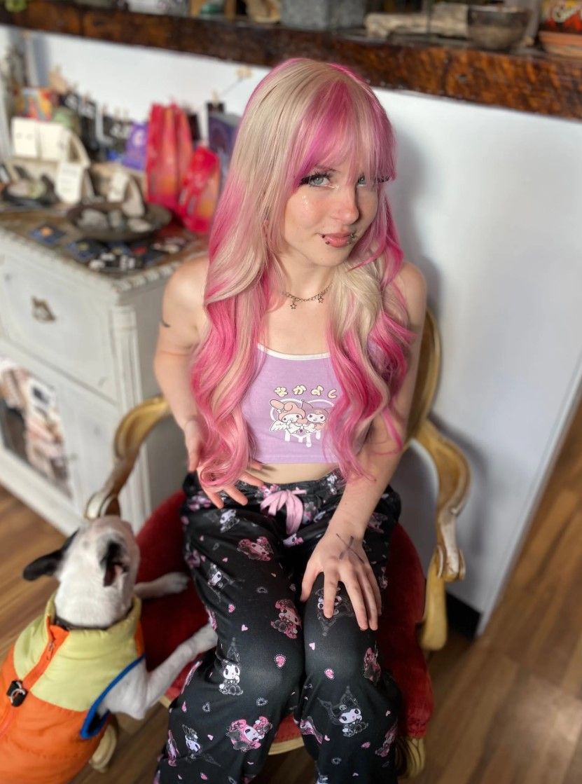 Woman with pink hair and dog sitting on a chair. She's wearing a crop top and patterned pants. The dog has a harness.