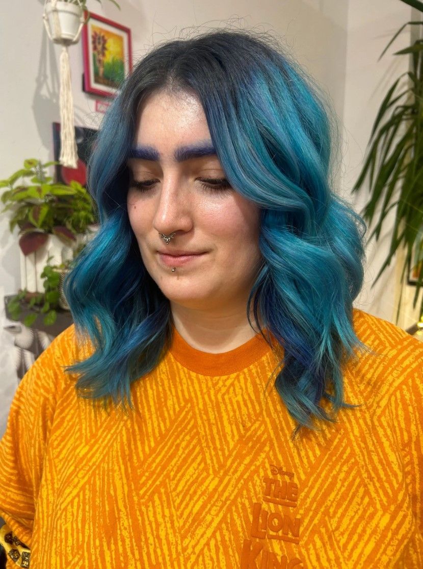 Woman with teal-blue wavy hair, wearing an orange patterned shirt, looking down. Indoor setting with plants.