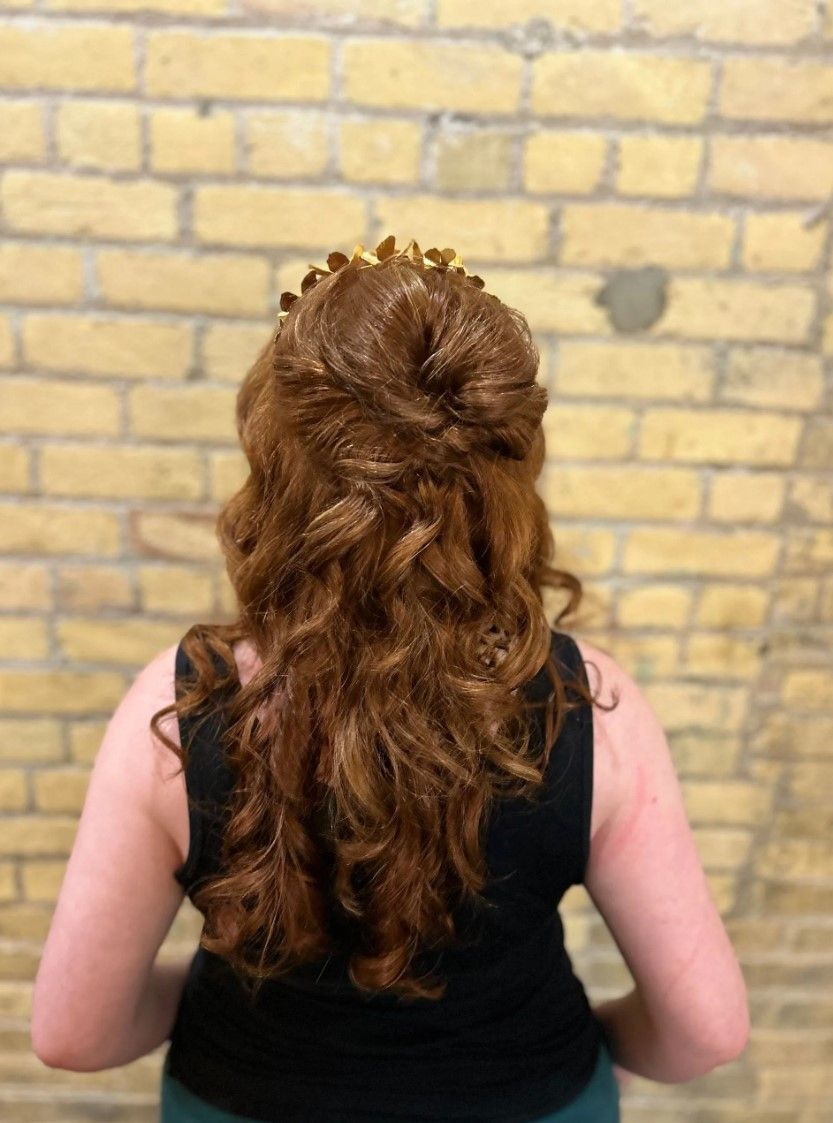 Woman with red wavy hair styled up and adorned with gold headpiece; stands in front of a brick wall.