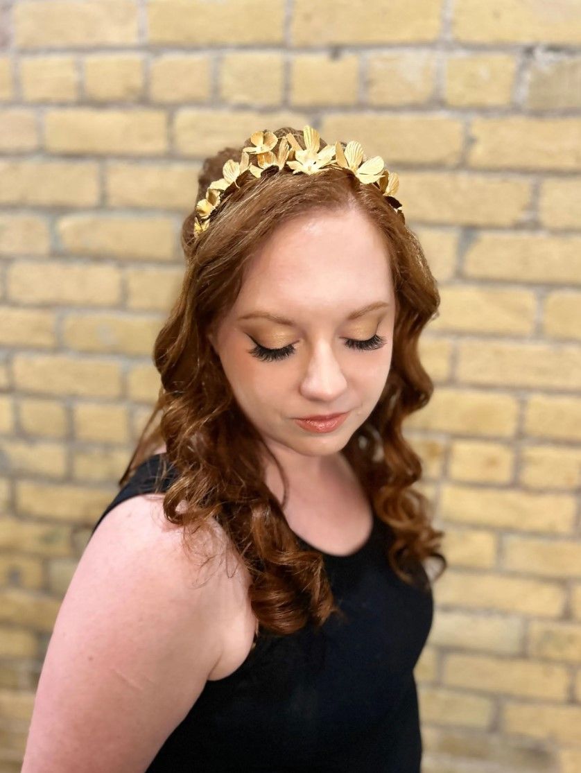 Woman with red hair, wearing a gold leaf crown, in front of a brick wall.