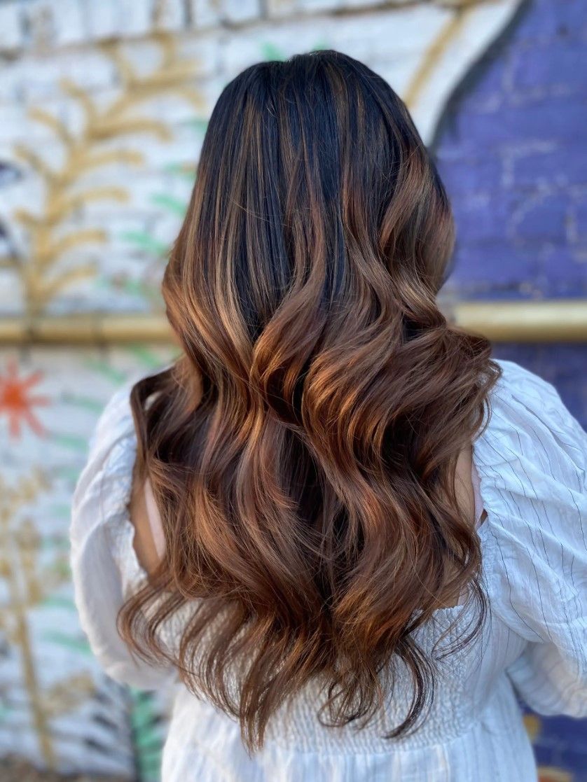 Woman with long, wavy brown hair with caramel highlights, wearing a white blouse, against a mural.