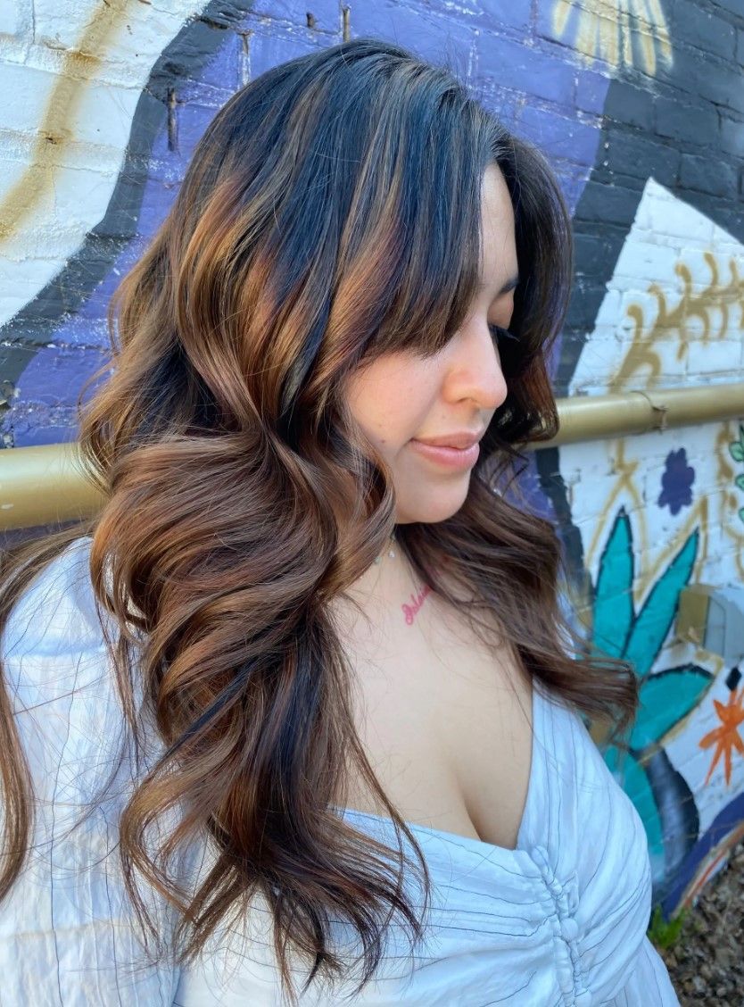 Woman with long wavy brown hair, looking down. Wearing a light-colored top, standing in front of graffiti.