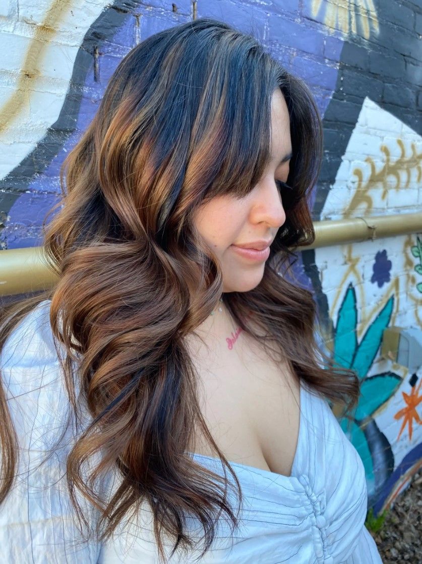 Woman with wavy, dark hair, wearing a light blue top, posing outdoors with a mural background.