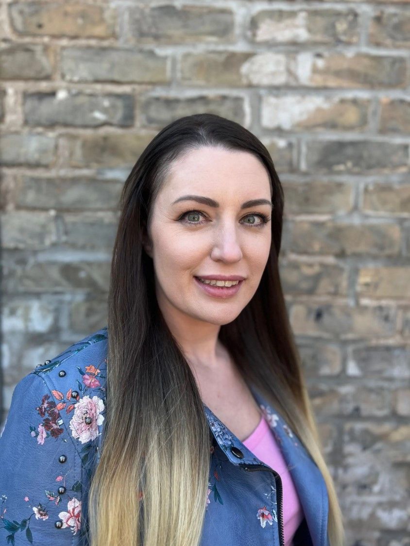 Woman with long hair smiles, wearing a pink shirt and blue jacket with floral details, brick wall backdrop.