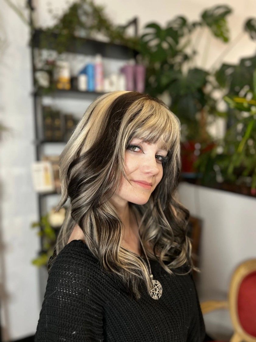 Woman with long, wavy hair, dark brown and blonde streaks, smiling. In front of a shelf with products and plants.
