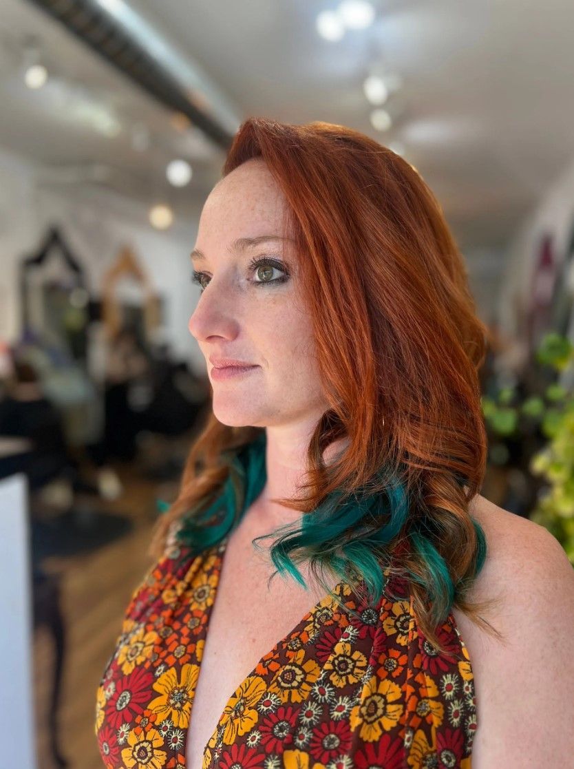 Woman with red hair and teal tips, in a floral print dress, at a salon.