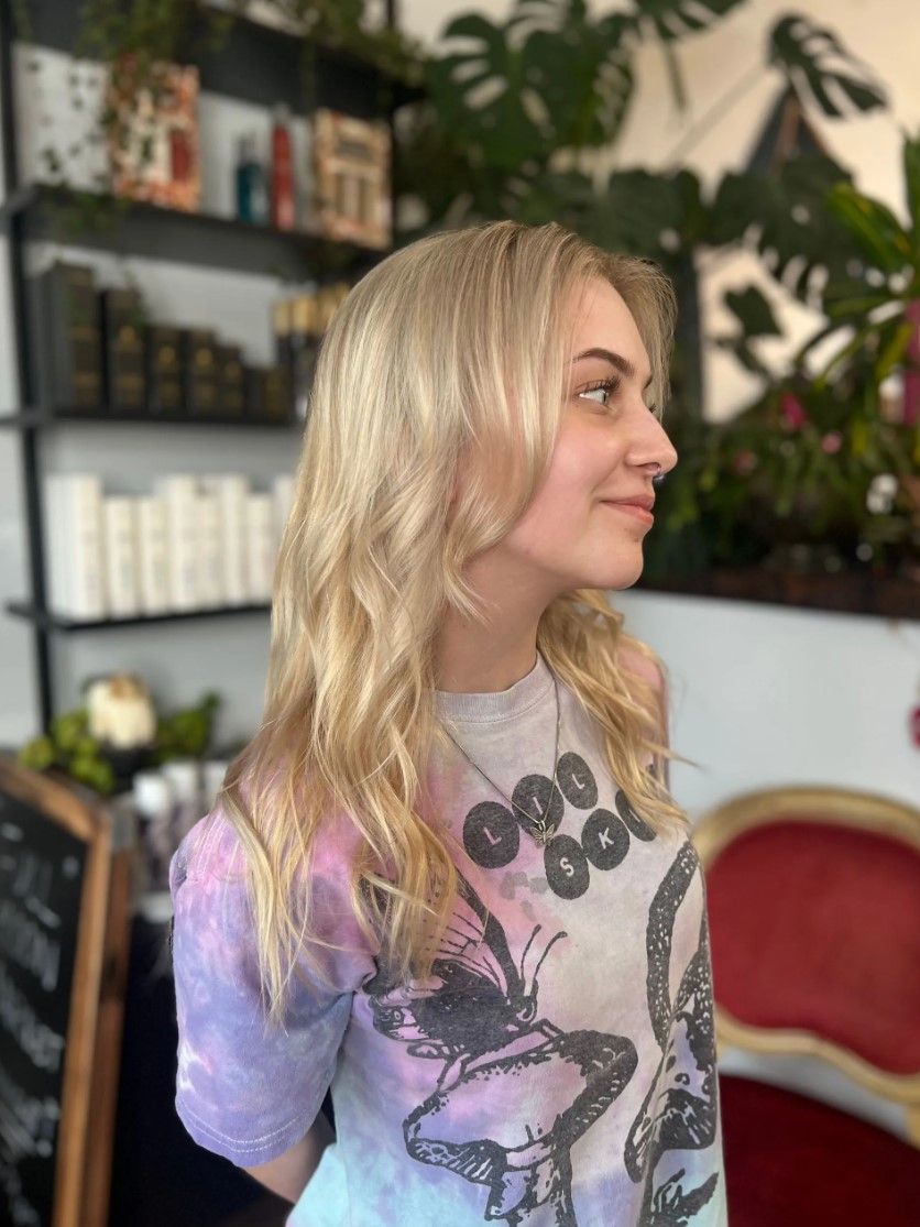 Blonde hair woman smiles, in a salon. Wearing a tie-dye shirt with a pendant.