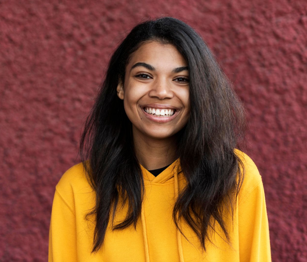 Woman wearing a yellow hooded sweatshirt smiles in front of a textured red wall.