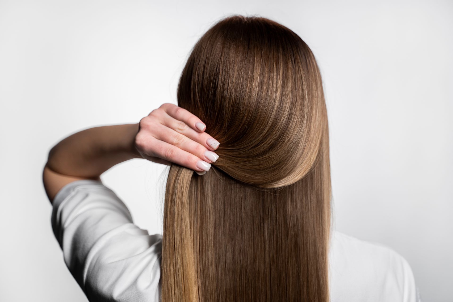 Woman with long, straight brown hair, holding a section up near the back of her head, against a white backdrop.