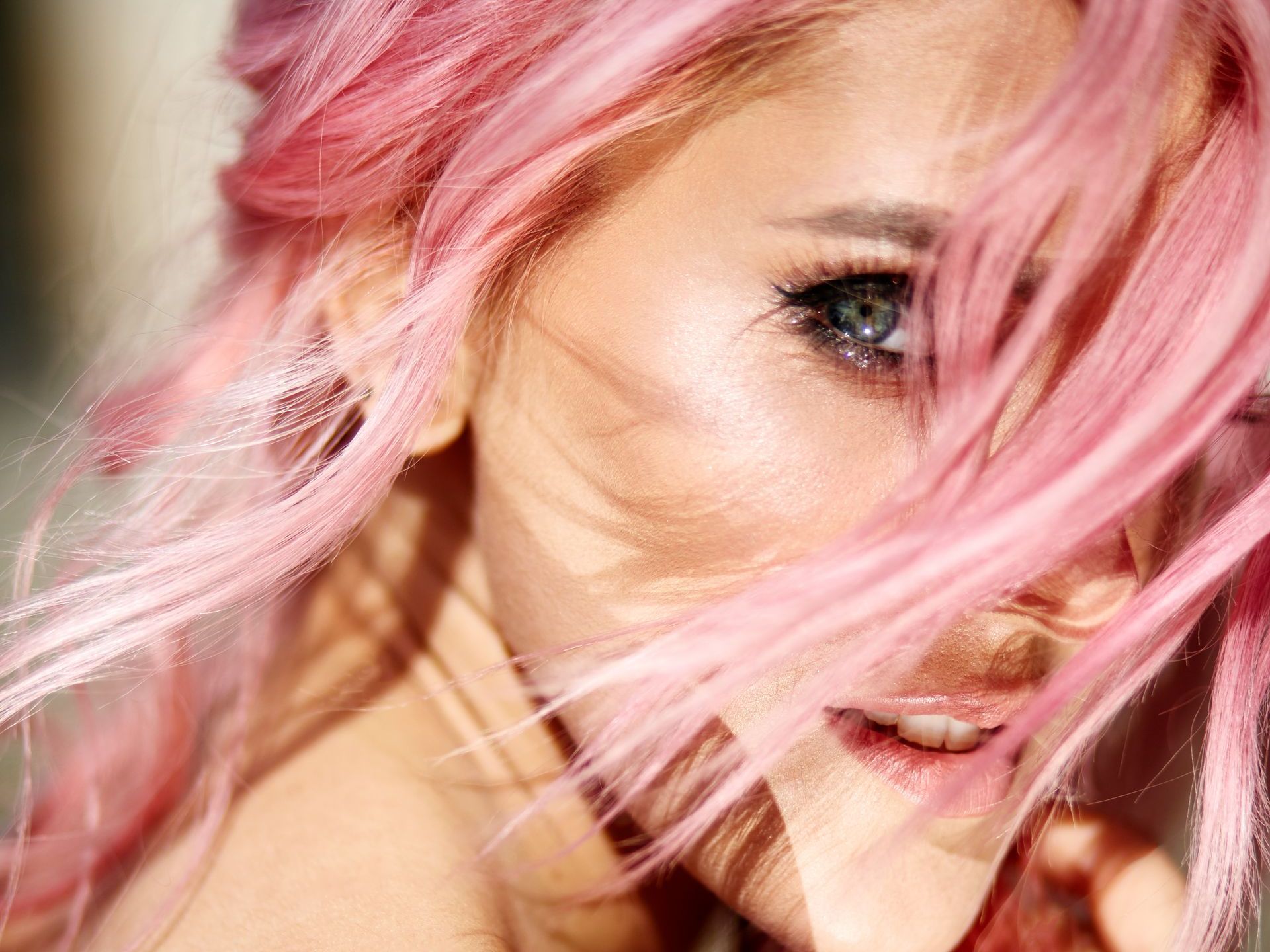 Woman with pink hair, blue eyes, smiling, hair partially covering face.